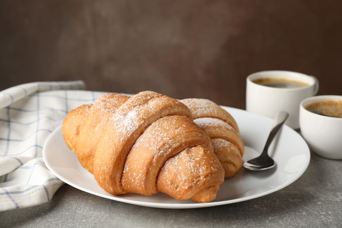 Two croissants with powdered sugar on plate with coffee on grey table, close up