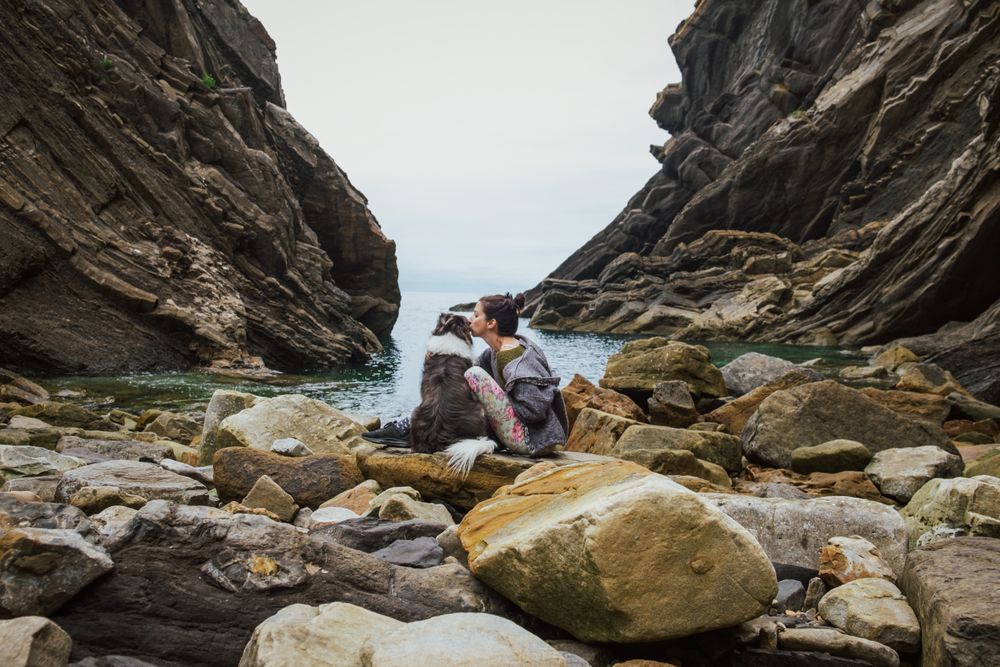 Traveling woman kissing dog sitting on boulder coast