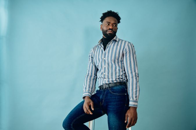 Black male model posing in blue studio in jeans and striped shirt leaning on step ladder