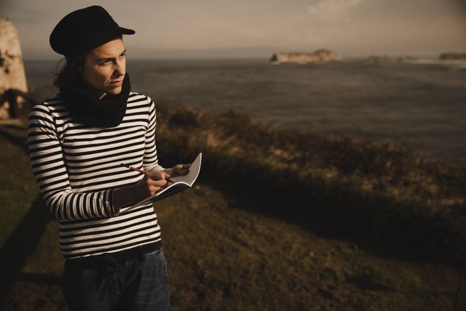 Woman writing in notebook on bench on shore near water