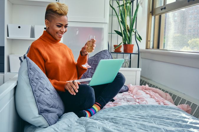 Happy Black woman smiling at a laptop with champagne in hand in a bright modern living room