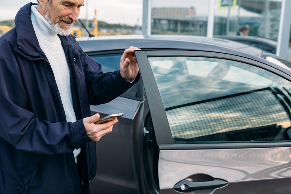 Man with grey beard opening a door of his car while looking at his phone
