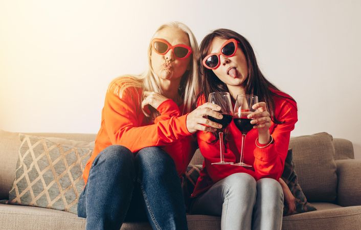 Women in similar outfits and fancy eyewear toasting wine and making faces sitting on couch