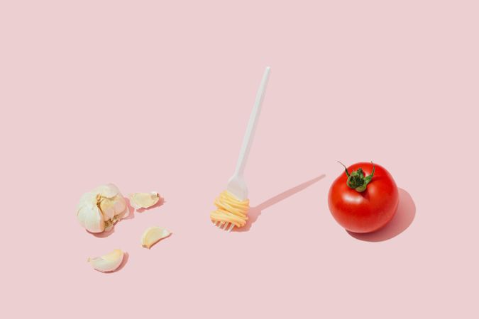 Garlic bulb, plastic fork with pasta, and whole tomato against pastel pink background