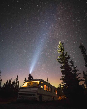 Silhouette of person sitting on top of camping van parked beside green trees under starry night