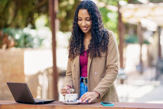 A young woman enjoys coffee while working on her laptop in a cozy outdoor setting