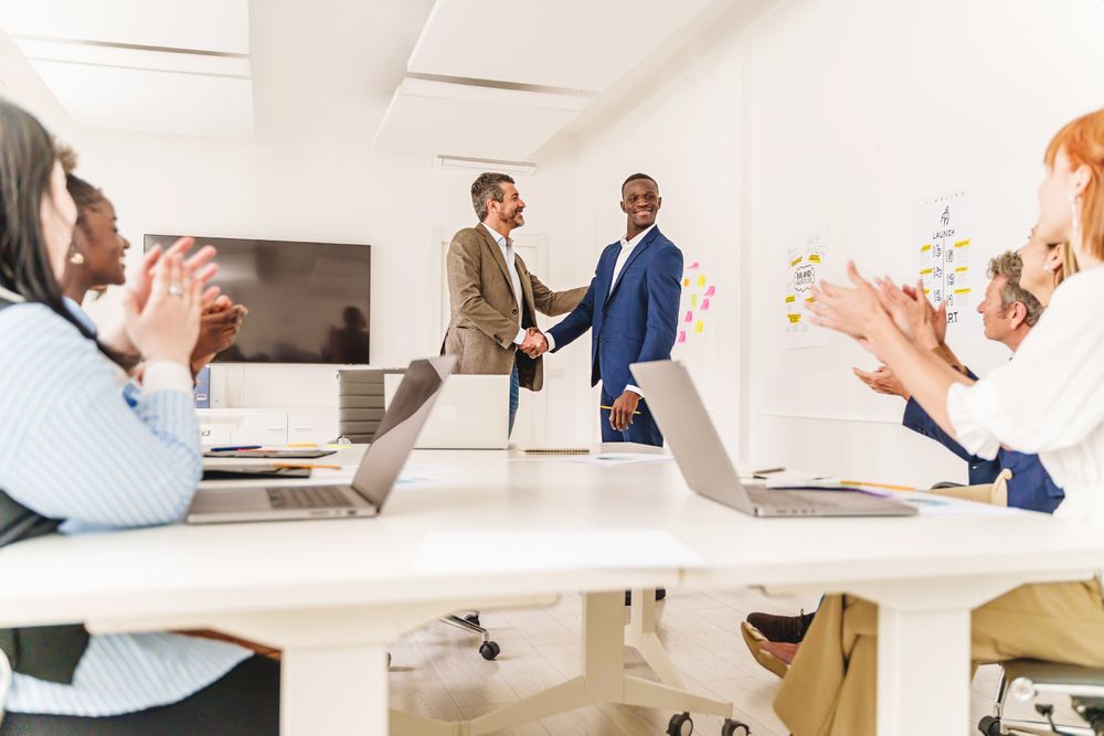 Wide shot of a diverse office with two people shaking hands at the center, while colleagues applaud deal being made