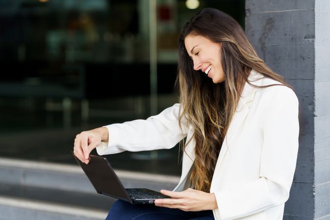 A Smiling Woman is Working on Her Laptop Outdoors While Dressed in Casual Business Attire