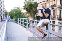 Young male with blonde bangs sitting on railing while using a mobile phone 5XrYBM
