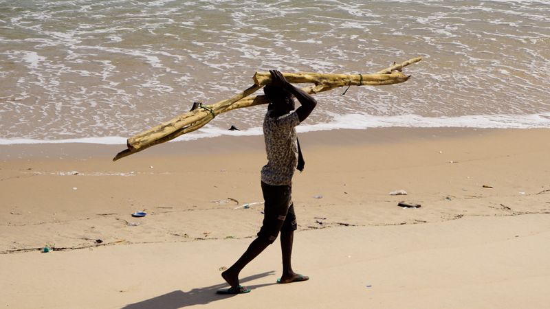 Side view of teenage boy holding a bunch of tree branches walking on seashore