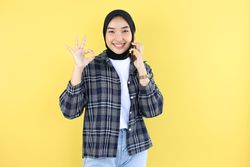 Young Asian Woman Wearing Hijab Talking on Cell Phone with Friend, Looking at Camera While Posing Okay.