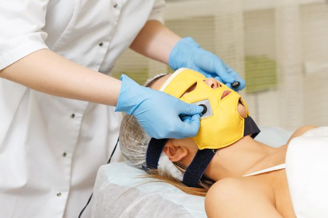 Female beautician placing yellow mask on woman’s face during facial