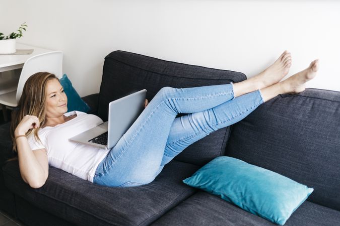 Relaxed long haired female lying on sofa with laptop computer working remotely from home