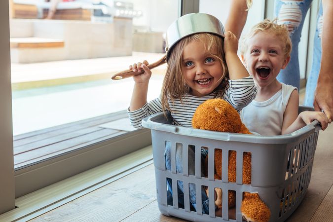 girl wearing a bowl as helmet with boy laughing while sitting in laundry basket