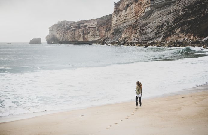 Woman walking on beach in green puffer jacket on fall day with footprints in sand