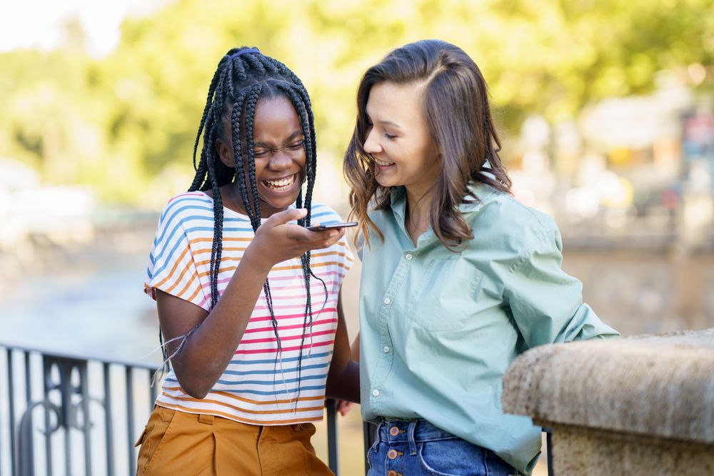 Female friends chatting on speaker phone in casual clothing outside