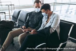 Couple with face masks sitting at airport and using mobile phone to check flight information 4AyBN4