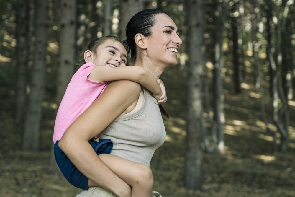 Happy mother giving her smiling daughter a piggyback ride, family having fun in the forest.