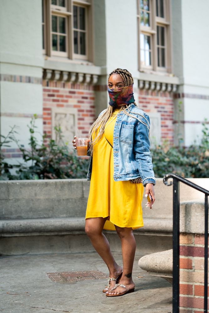 Woman in yellow dress and gator mask standing in front of classic building holding iced coffee