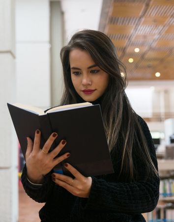 Woman standing along bookshelves and holding open book