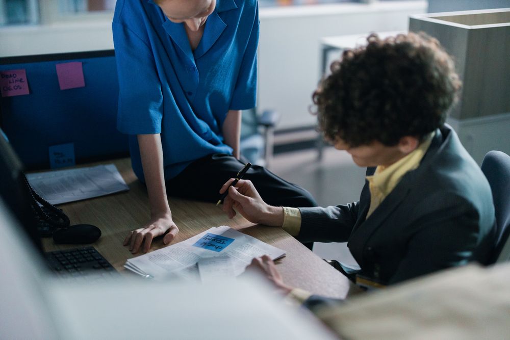 Smiling colleague talking to another colleague who is writing on document on her desk