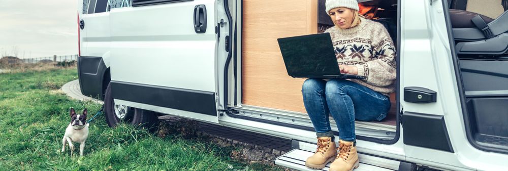 Female in sweater working remotely sitting on step of van while on a road trip, wide