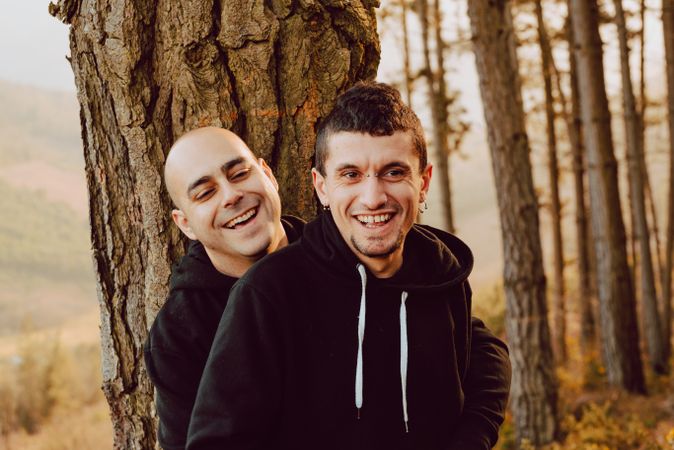Smiling men hugging near tree in forest on hill