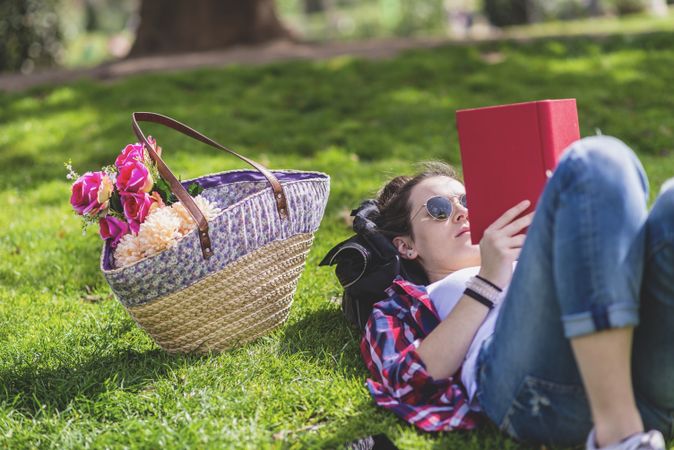 Side view of a hipster happy woman lying on grass in sunny day at park while reading a red book
