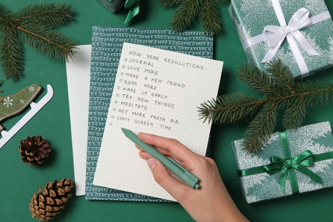 Paper with list, pen in hand, fir branches and gift boxes on green background, top view