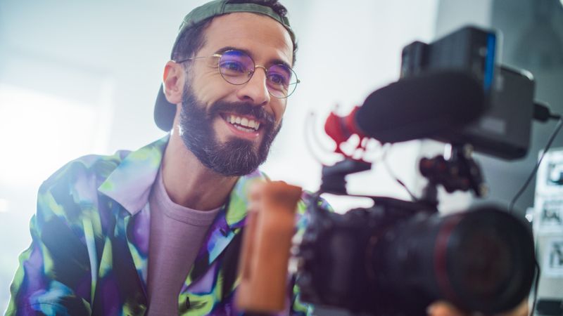 Smiling Young Latin Male Filmmaker Operating A Professional Camera In A Well-lit Studio, Capturing Engaging Content For A Modern Advertising Or Movie Production Project.
