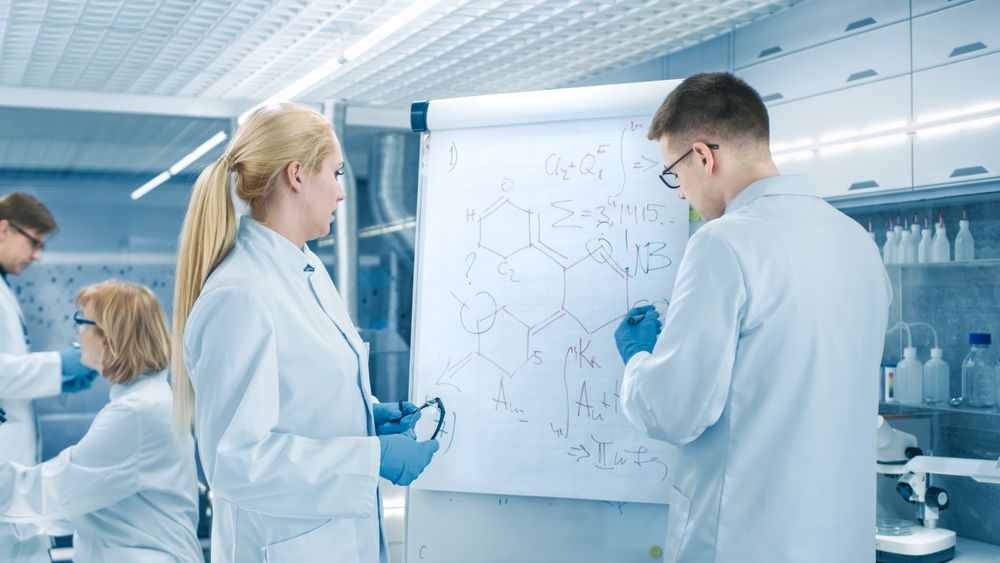 Young Female and Male Scientists Write Formulas on a Whiteboard.