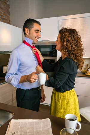 Curly haired woman adjusting tie of smiling businessman while having fast breakfast before work