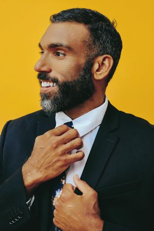 Happy Black male in suit adjusting his floral tie and looking away in yellow studio