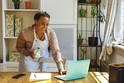 Female entrepreneur in work apron taking video meeting on laptop in bright office