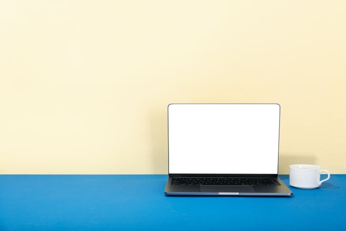Open laptop with blank mockup screen in cream and blue with cup and saucer with copy space