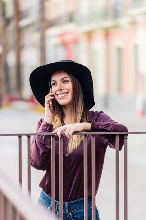 Charming woman in trendy outfit talking on smartphone and smiling at city street