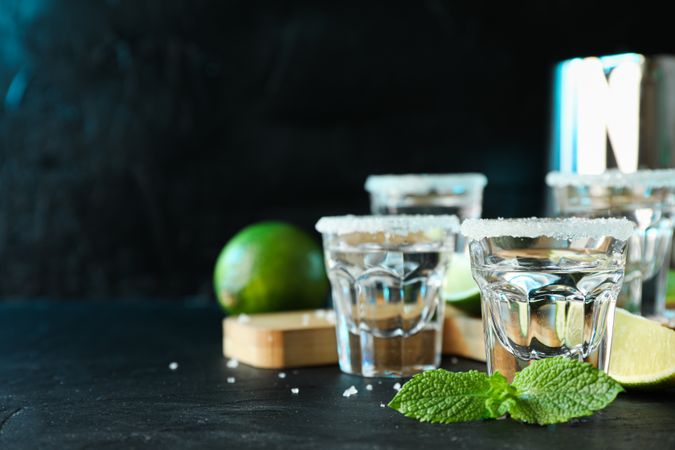Tequila shots with salt, lime slices and mint on dark table