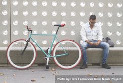 Male checking phone while sitting with bike parked in front of patterned cement wall bE2YG0