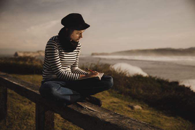 Woman writing in notebook and sitting on bench on shore near water