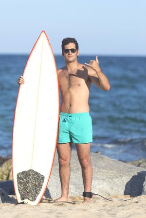 Male surfer standing with red outlined board making the “hang loose” sign