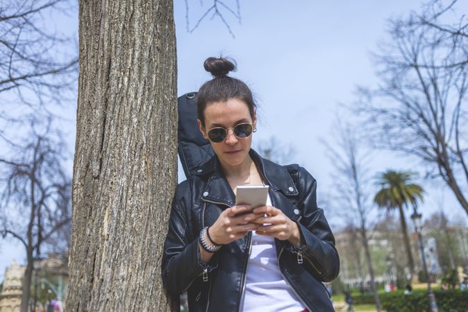 Side view of a young laughing hipster woman standing and leaning on a tree at park in sunny day while using a mobile phone