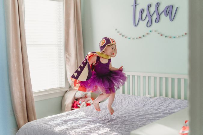 Girl in a cape and purple dress jumping on bed during daytime