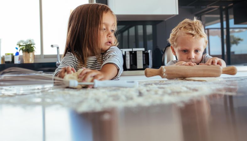Little boy using rolling pin and girl kneading dough with flour on kitchen table