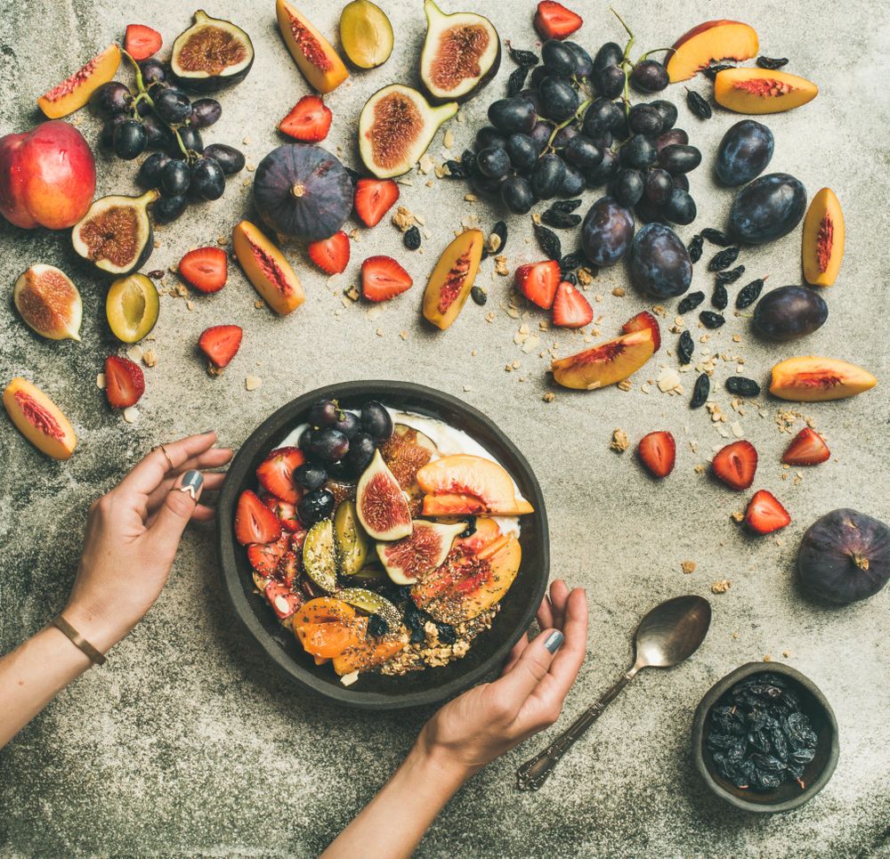 Woman holding bowl of yogurt over table of figs, peaches, and chia seeds, square crop