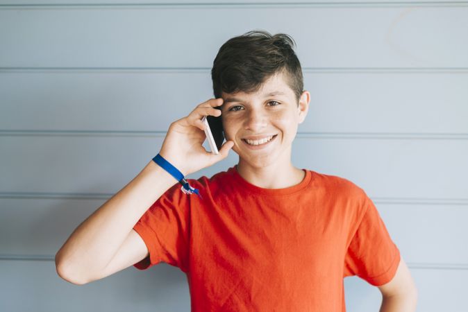 Smiling teenager in red t-shirt standing against wall while using phone