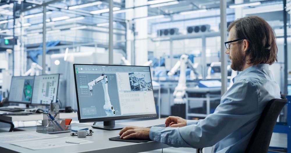 Portrait of a Young Factory Manager Working on Desktop Computer in Industrial Manufacture with Autonomous Assembly Robots. Production Line Making Heavy Machinery Parts in the Background