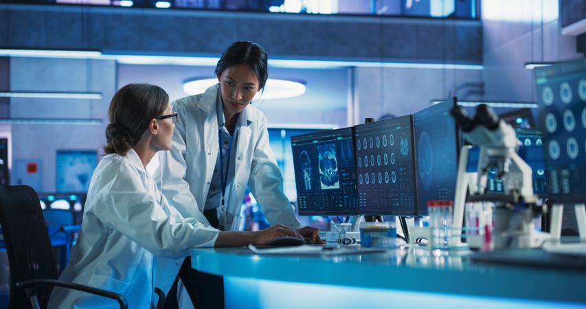 Asian And White Female Doctors Talking In Medical Research Center And Using Desktop Computer To Analyze MRI Scans Of Brain.