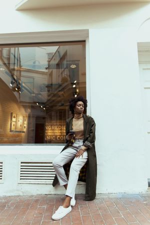 Black woman in green jacket holding a cup of coffee leaning on wall beside a store