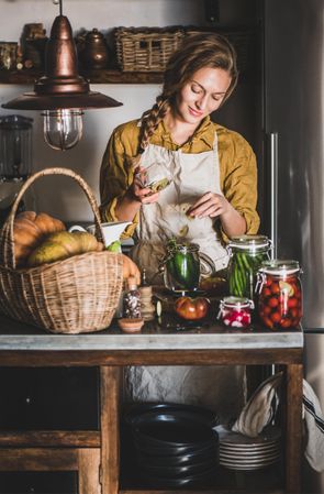 Young woman in rustic kitchen smiling putting herbs into jar of cucumbers for pickling