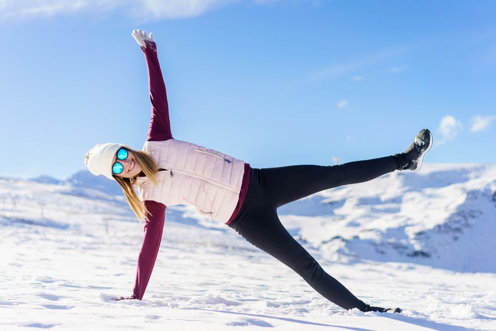 Woman in snowsuit doing side plank with leg up on snowy mountain